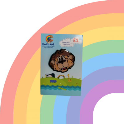 ID: White background with a faded rainbow. Noah's Ark Charity lion badge in the forefront on blue and green backing card.